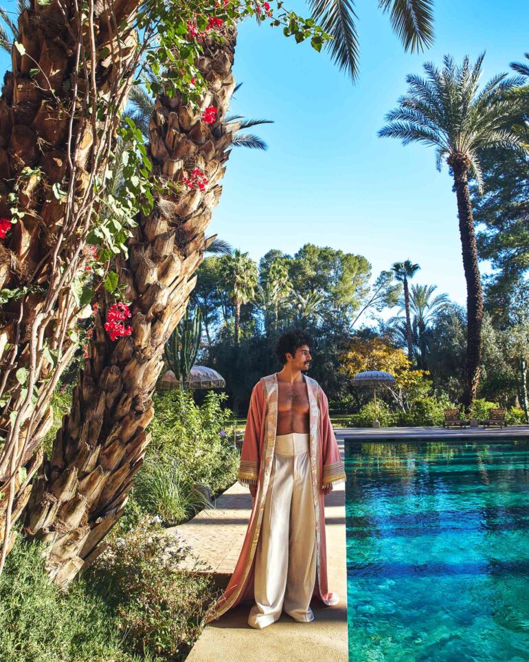Amran Bennaji wears a pink embroidered canvas Hilal kimono while looking out over a pool surrounded by palm trees in Marrakech, Morocco