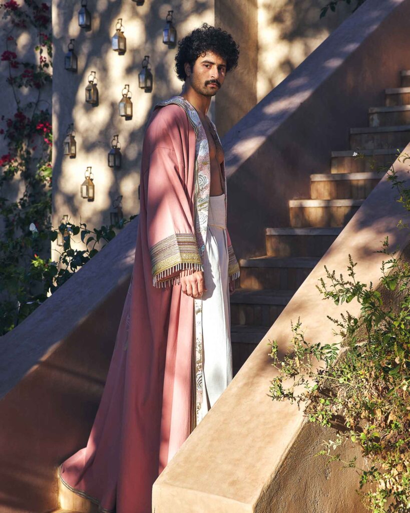 Amran Bennaji wears a pink embroidered canvas Hilal kimono while standing on stairs and in front of Moroccan lamps on a wall in Marrakech, Morocco