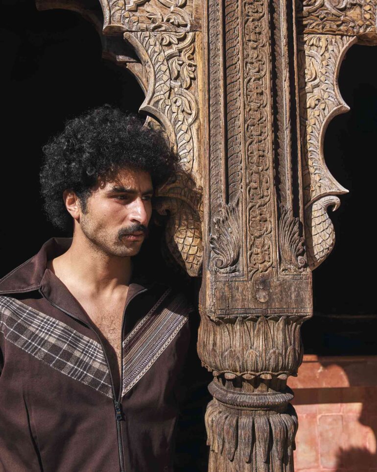 Amran Bennaji wearing a brown unisex jacket while leaning against an ornately carved wooden doorframe in Marrakech, Morocco