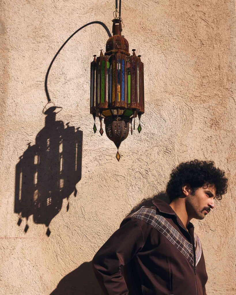 Amran Bennaji wearing a brown unisex jacket while standing next to a Moroccan lamp which casts a shadw onto a wall in Marrakech, Morocco