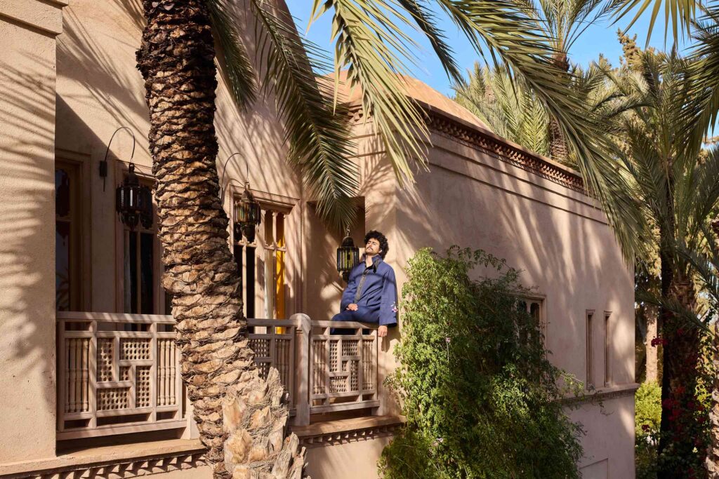 Amran Bennaji wears a navy 'Nomad' coat while sitting on the railing of a balcony surrounded by palm trees in Marrakech, Morocco