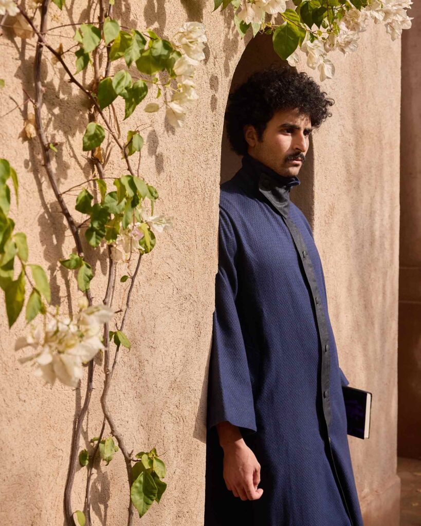 Amran Bennaji wears a navy 'Nomad' coat while posing next to flowers growing on a beige wall in Marrakech, Morocco