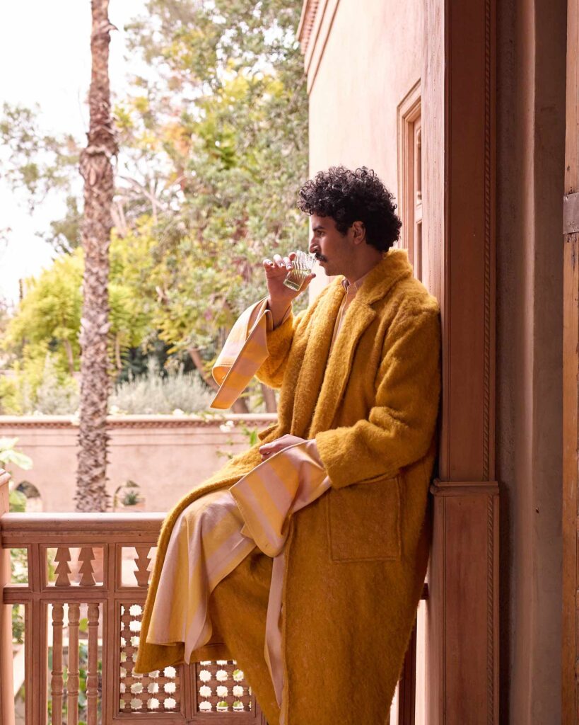 Amran Bennaji wearing a yellow blanket coat by Marrekeshi Life while sipping on a tea and looking out over a landscape from a balcony in Marrakech, Morocco
