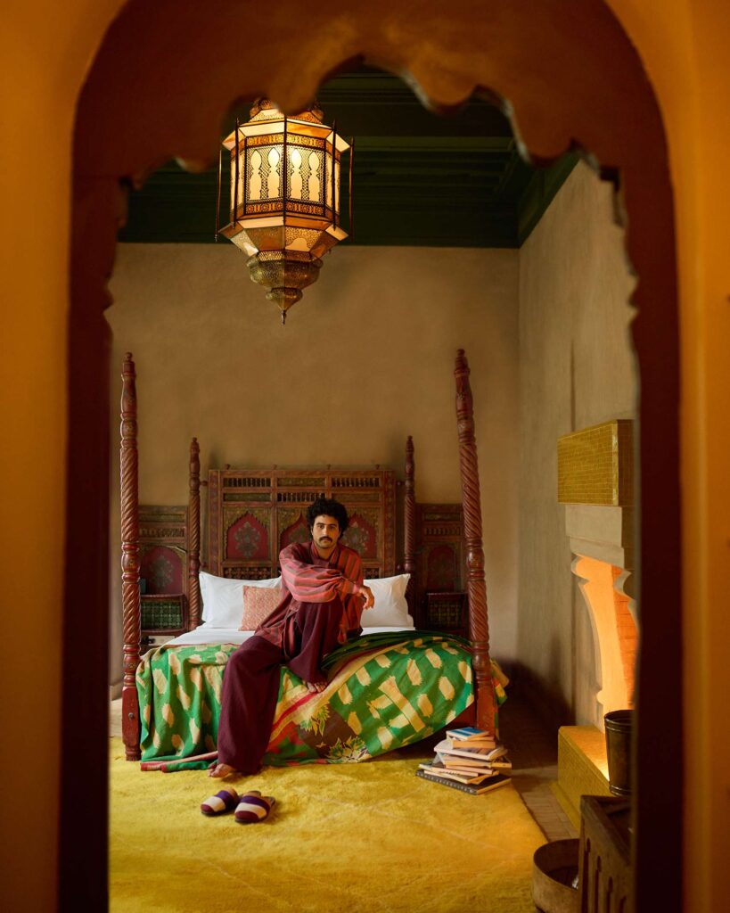 Amran Bennaji sits on a wooden four-poster bed with a large lamp hanging above in Marrakech, Morocco