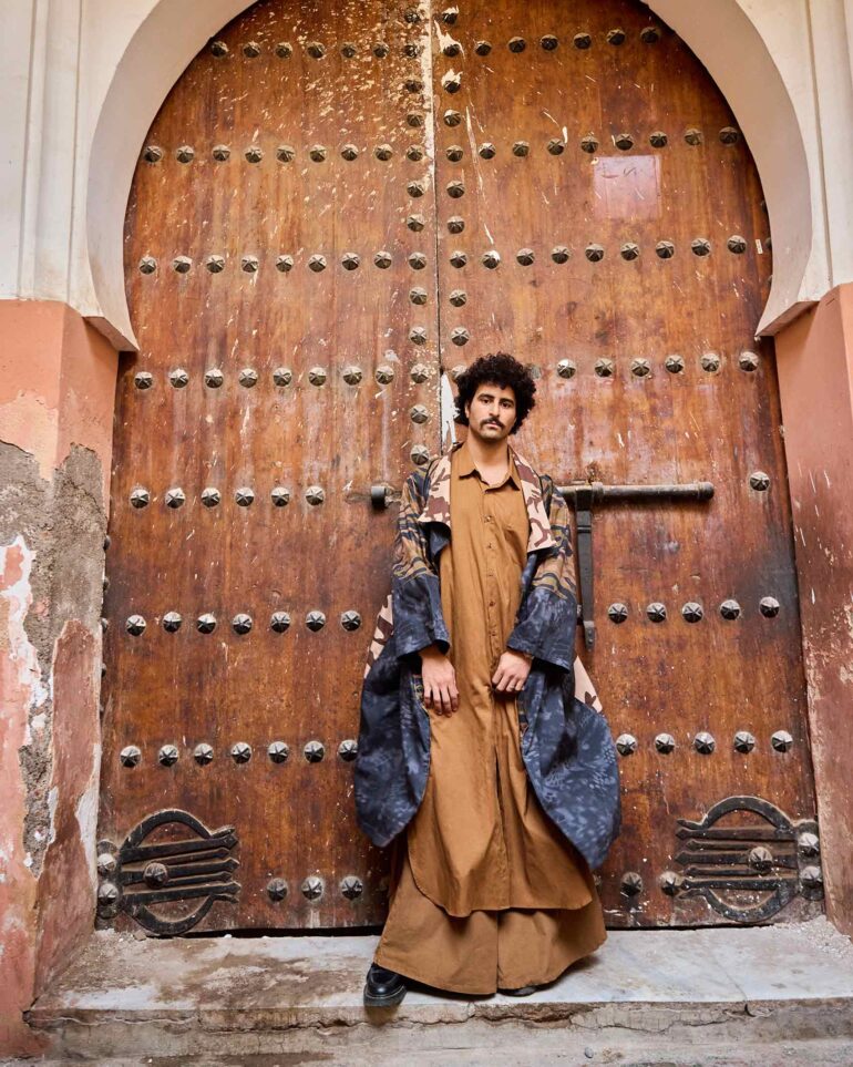 Amran Bennaji wearing a long, buttoned shirt while posing in front of a wooden doorway in Marrakech, Morocco