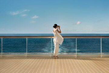 Woman wearing a flowing white sleeveless top and white trousers tiptoes on the deck of an Explora Journeys ship and looks through her binoculars out at the limitless horizon