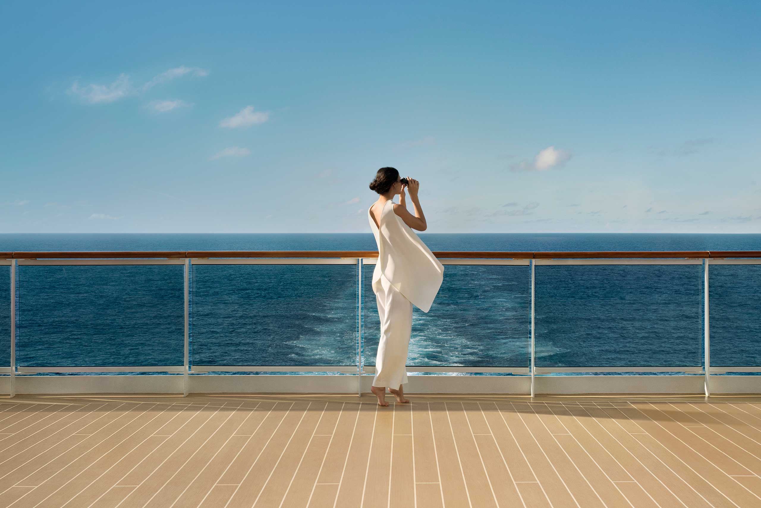 Woman wearing a flowing white sleeveless top and white trousers tiptoes on the deck of an Explora Journeys ship and looks through her binoculars out at the limitless horizon