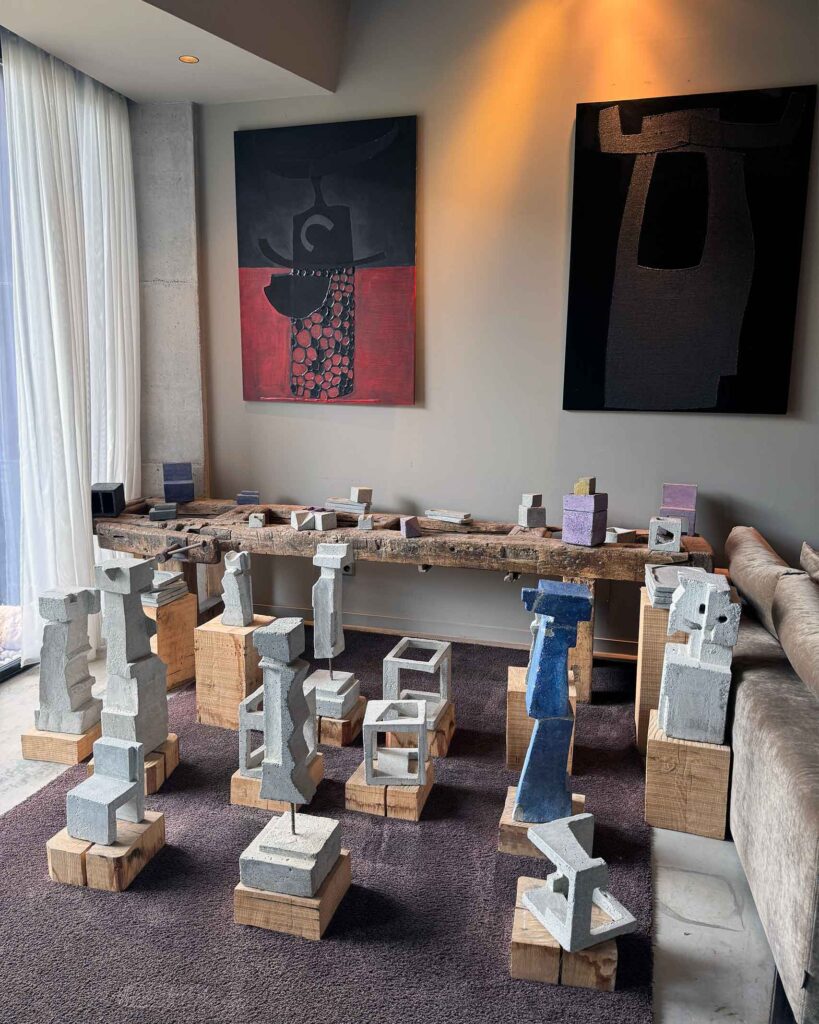 Art installation with abstract concrete sculptures displayed on wooden blocks beneath contemporary paintings.
