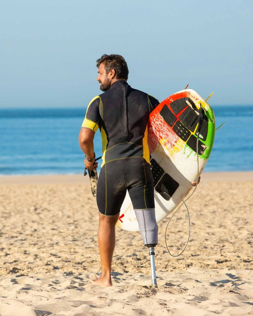 South Asian man with dark hair and prosthetic leg, holds a surf-board and wears a black and yellow wet-suit as he looks to sea