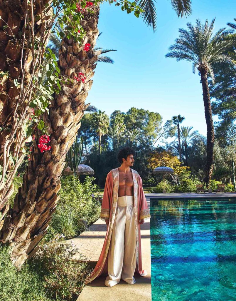 Model Amran Bennaji poses by the pool at Jnane Rumi in Marrakech. He wears a long, pink robe, bare chest, white corset and flowing white trousers. Behind him, the lush gardens of the hotel