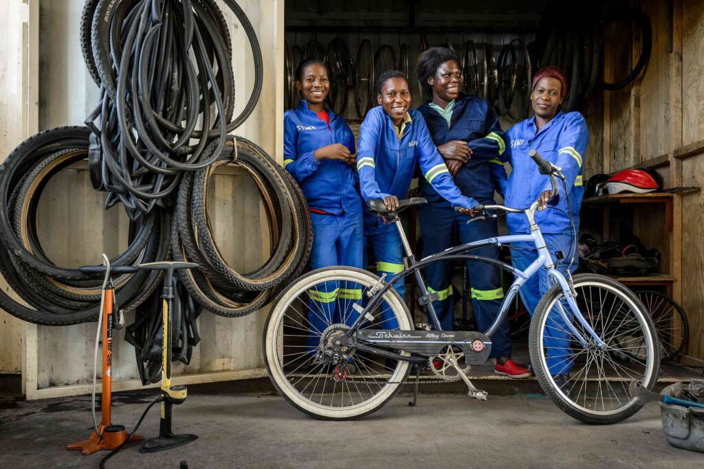 Four African women in blue tracksuits at the Chipego Bike Shop, poses with a re-engineered bicycle