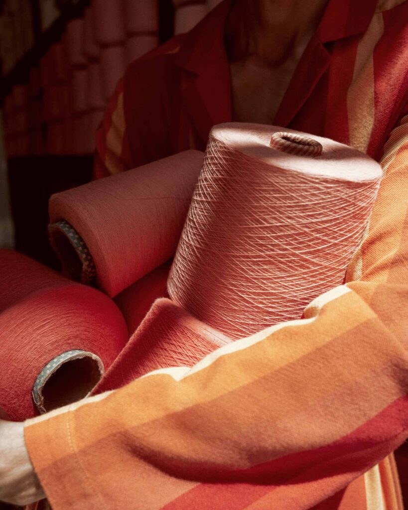 Close up shot of a man in orange and umber robe carrying spools of terracotta and peach toned thread