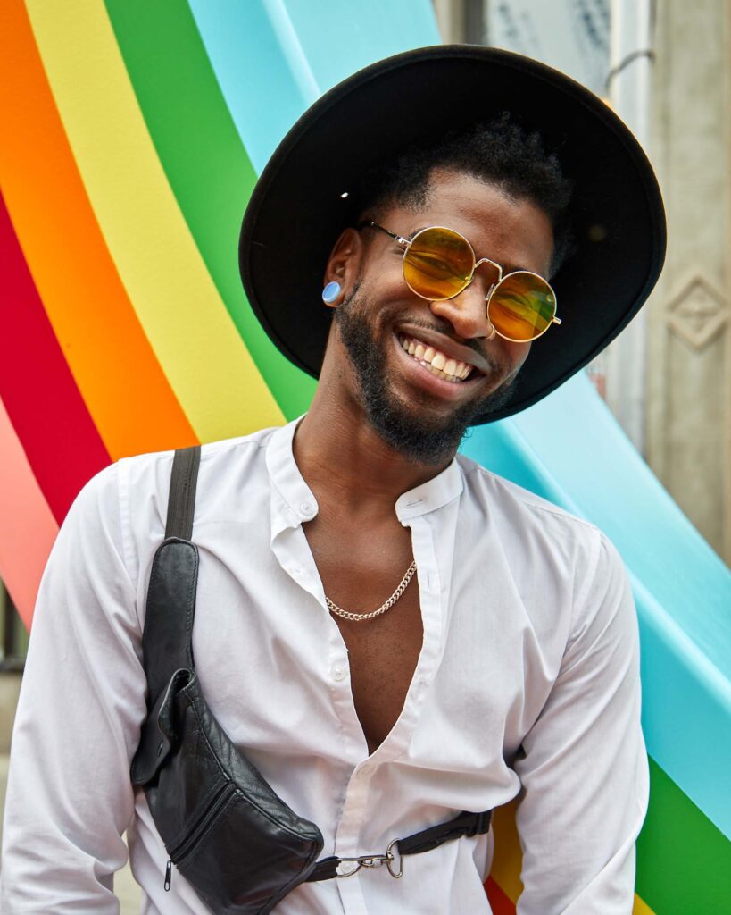 A black man smiles as he stands in front of a rainbow striped backdrop. He wears a black hat, has a beard, blue ear piercing, yellow tinted sunglasses, a nose-ring and a white collarless shirt open to his chest