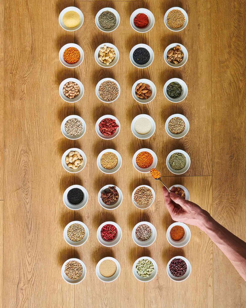 Spices and seeds in white saucers arranged 4 across and 8 down, while a hand appears from the right and scoops some onto a spoon. From the Awakened World issue