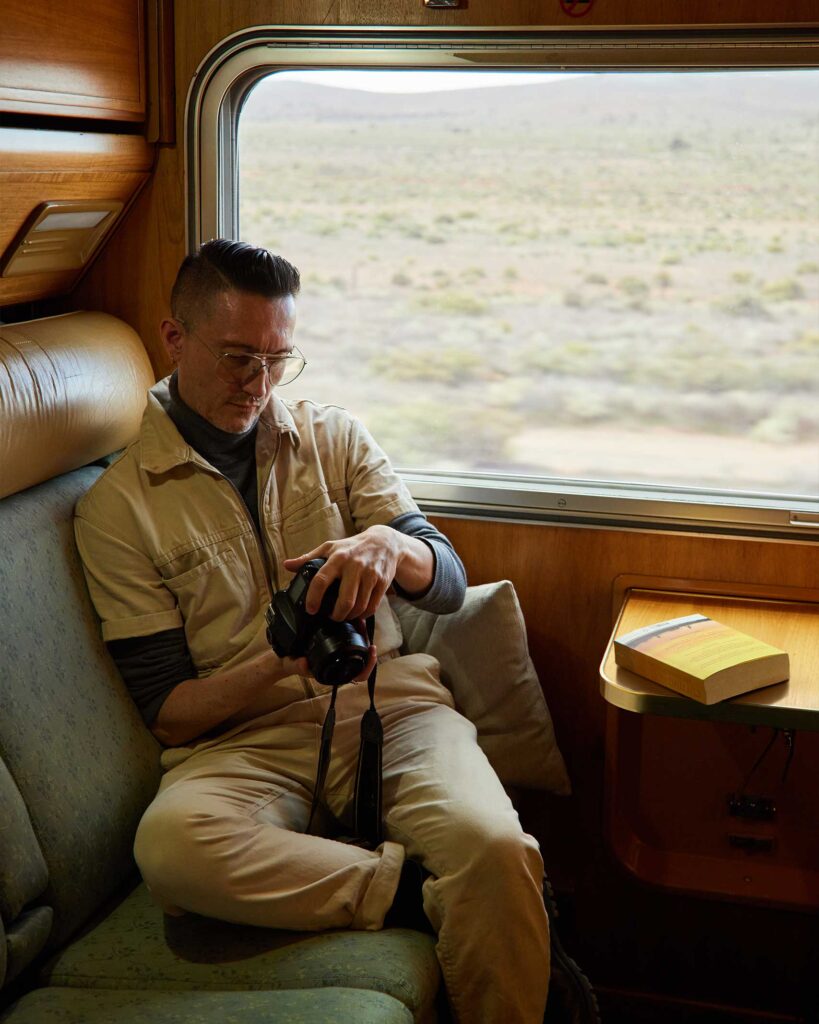 Martin Perry of OutThere wears a beige jumpsuit and sits by the window and wood panelled interiors of the Indian Pacific train in Australia. he look at an image taken on the back of his SLR camera. From the Awakened World issue