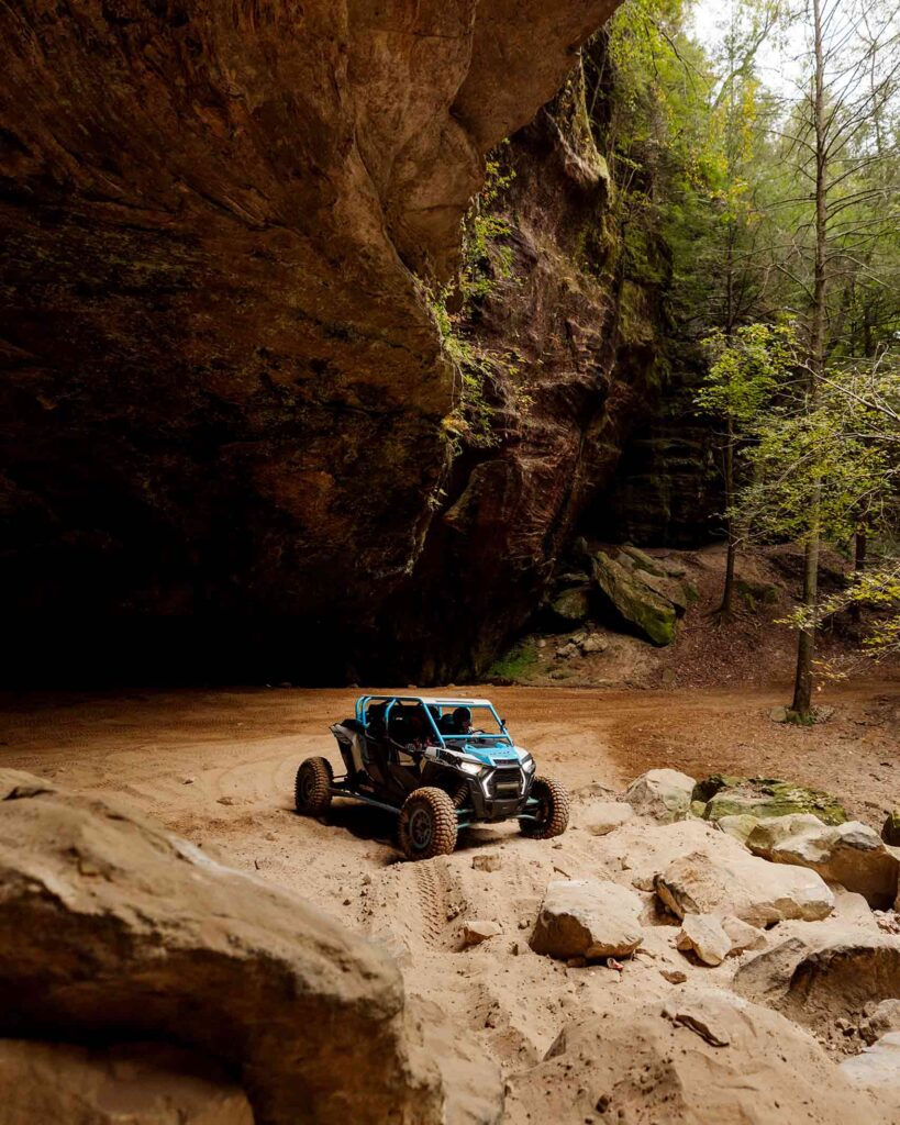 A blue off-road ATV sits on a sandy bank underneath a cliff in a forest in Kentucky. From the Awakened World issue