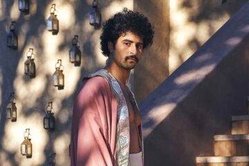 Amran Bennaji wears a pink embroidered canvas Hilal kimono. He has curly balck hair and a moustache. He stands in front of a wall full of small lanterns, with dappled sunlight