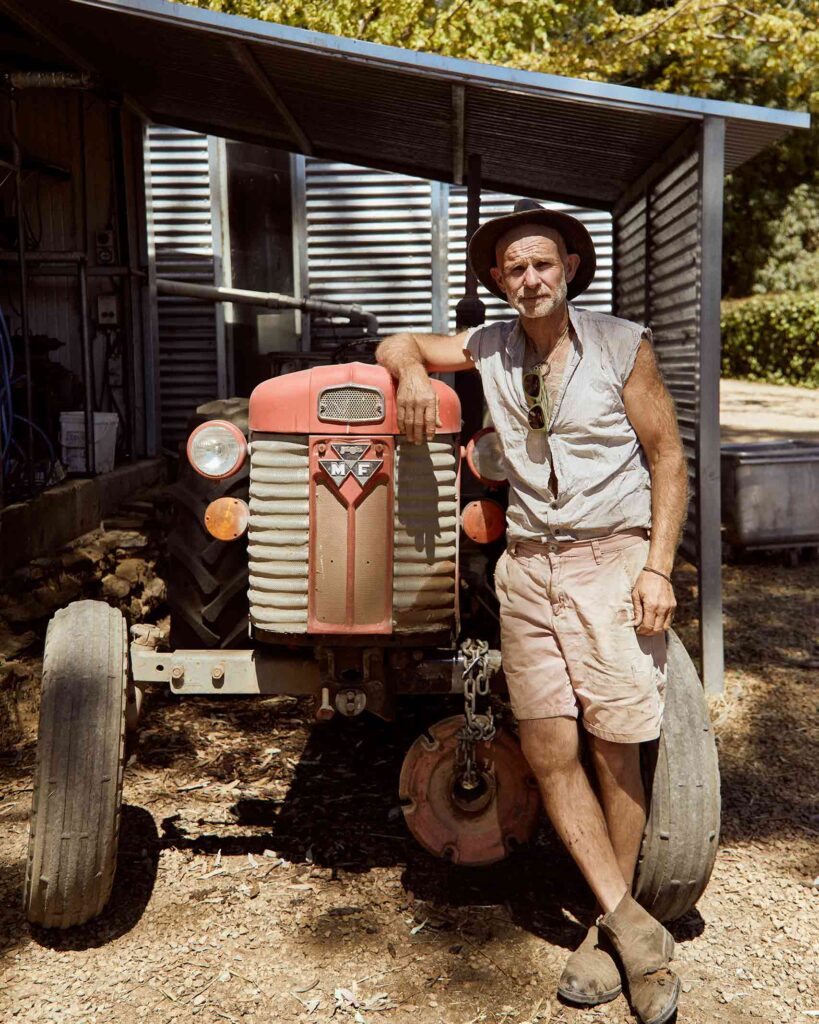 The Dairy Man Barossa, a white man wearing a brown hat, cut off linen shirt and beige shorts stands by a red tractor at his farmstead in the Barossa Valley. From the Awakened World issue