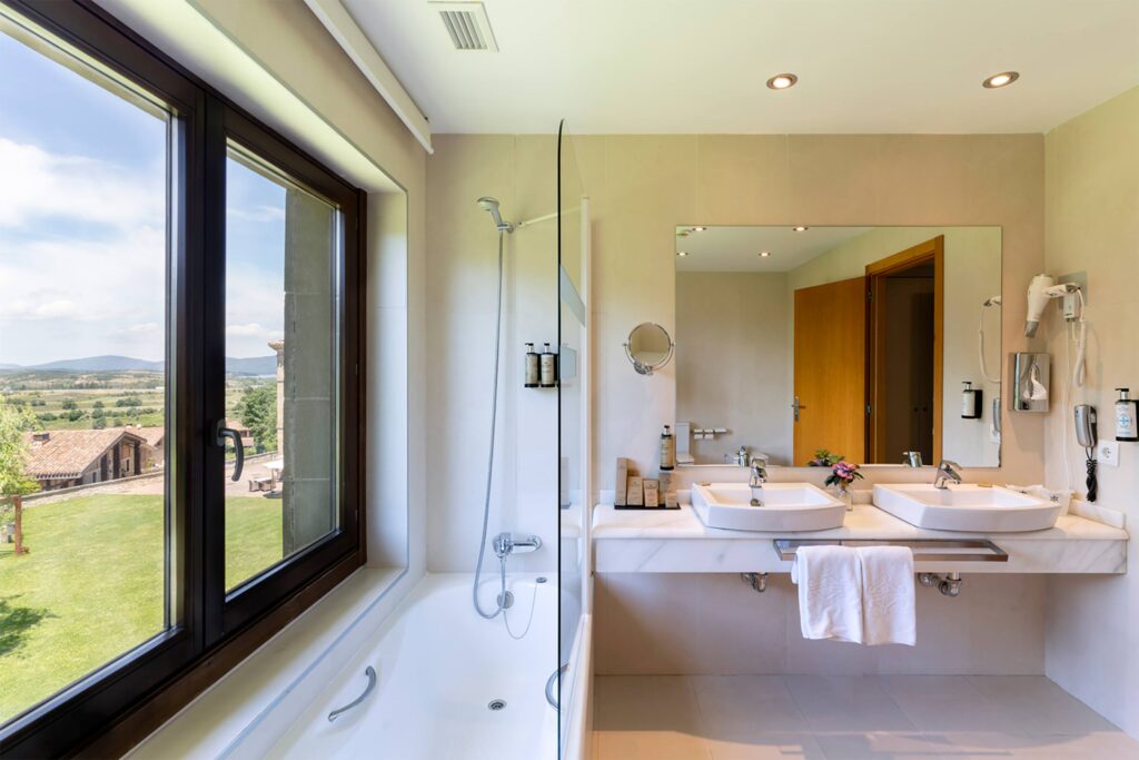 Modern hotel bathroom with double sinks, glass shower over bathtub, and a large window overlooking rolling Basque countryside.