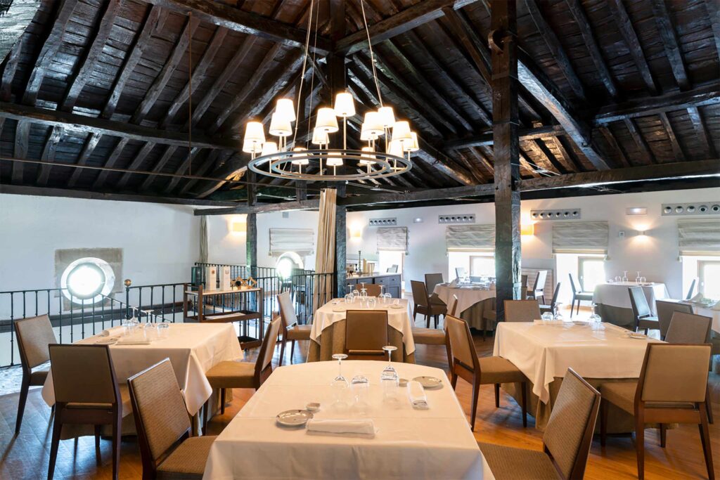 Elegant restaurant dining room at Parador de Argómaniz with exposed wooden beams, circular chandelier lighting, and tables neatly set with white tablecloths.