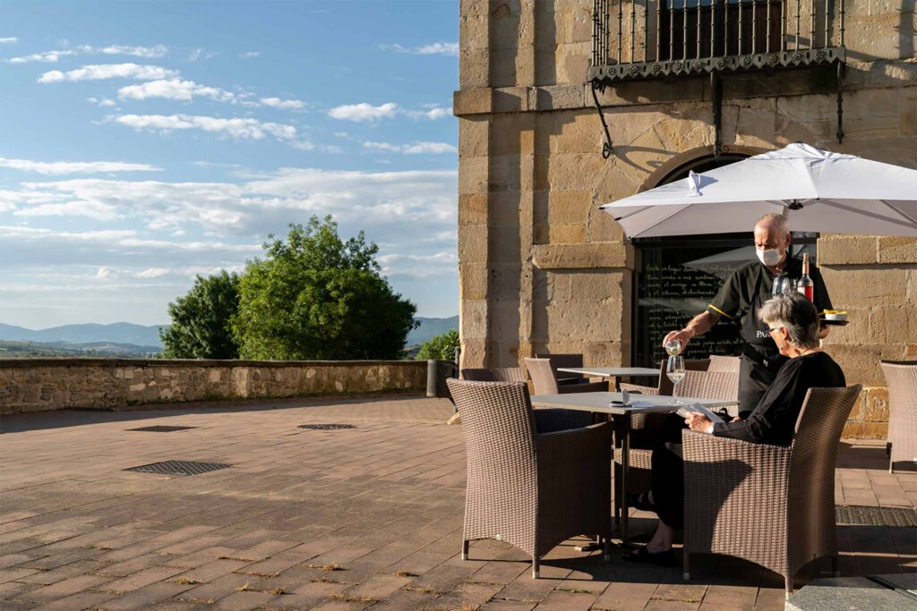 Outdoor terrace at a historic stone hotel where a waiter pours wine for a guest seated beneath a large umbrella.