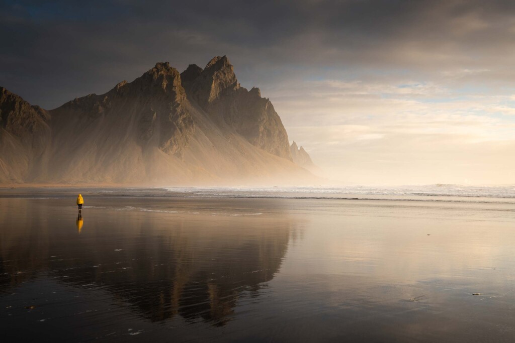 Person in bright yellow jacket walks on a black volcanic beach, flanked by dramatic mountains in the background in Iceland – summer travel with Scott Dunn