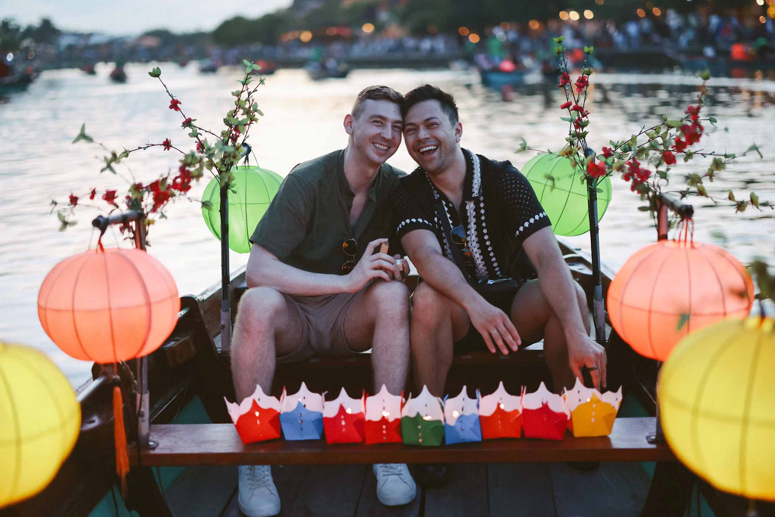 A male-male, both white with dark hair, one in a green shire and tan shorts, the other in a black, patterned shirt with dark shorts, sith on a lantern and flower adorned boat in Vietnam