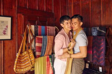 A male-male couple, both Filipino-Asian, embrace in the boutique shop – amidst a selection of Thai textiles – at the 137 Pillars House in Chiang Mai Thailand, a LGBTQIA2S+ friendly Small Luxury Hotels of the World property. They both have short dark hair, one wears a pink polka dot shirt, the other a light blue polo