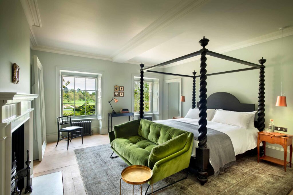 A black four-poster bed with a green sofa, a fire place and large sash windows with views of the gardens at The Newt in Somerset, Bruton, United Kingdom