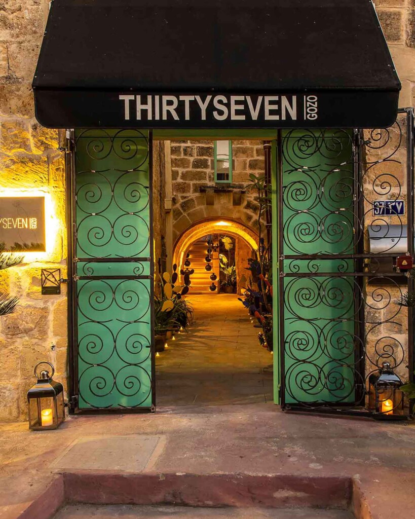Entrance to Thirtyseven Gozo at dusk, with a black awning spelling out its name and double green doors leading to a candlelit alleyway entrance