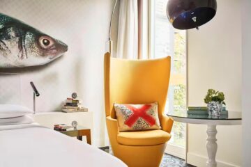 Room detail of the Andaz Amsterdam Prinsengracht showing a fish mural above a white bed and a bright mustard, sculptural armchair, with blue detail carpet and mid-century modern overhead glass lampshade