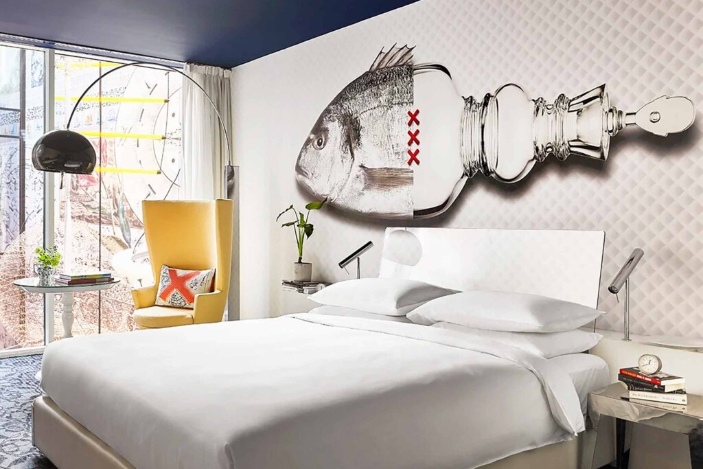 Room detail of the Andaz Amsterdam Prinsengracht showing a fish mural above a white bed and a bright mustard, sculptural armchair, with blue detail carpet and mid-century modern overhead glass lampshade