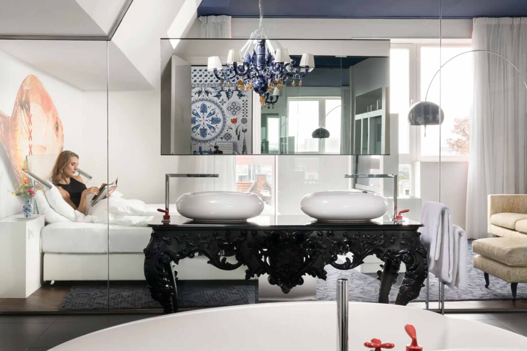 Bathroom detail of the Andaz Amsterdam Prinsengracht with open plan design. A woman lies reading in bed in the background, but in the foreground we see the bathroom, with twin vanity sinks, blue chandelier and bathtub with red taps