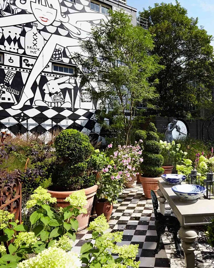 The sun trap garden of the Andaz Amsterdam Prinsengracht adorned with greeneary, set on a chequerboard tiled floor. In the background, on a wall spanning the entire building is a piece of monochrome street art depiction of a contemporary Alice in Wonderland story, complete with march hare and "drink me" bottle