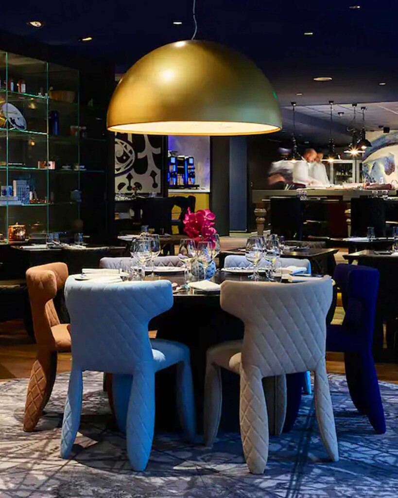 Dining area at Andaz Amsterdam Prinsengracht, with blue, grey and pink padded chairs surrounding a round black table with crockery and cutlery laid. Overhead, a large gold lampshade