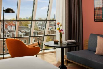 View from inside room at Avani Museum Quarter Amsterdam out over the rooftops of the city through chequered glass windows. IN front of the window, an orange armchair with an AvaniFit yoga mat on it, as well as a table with tulips, macaroons and photography artbooks, next to a grey mid-century modern sofa set against a teracotta wall