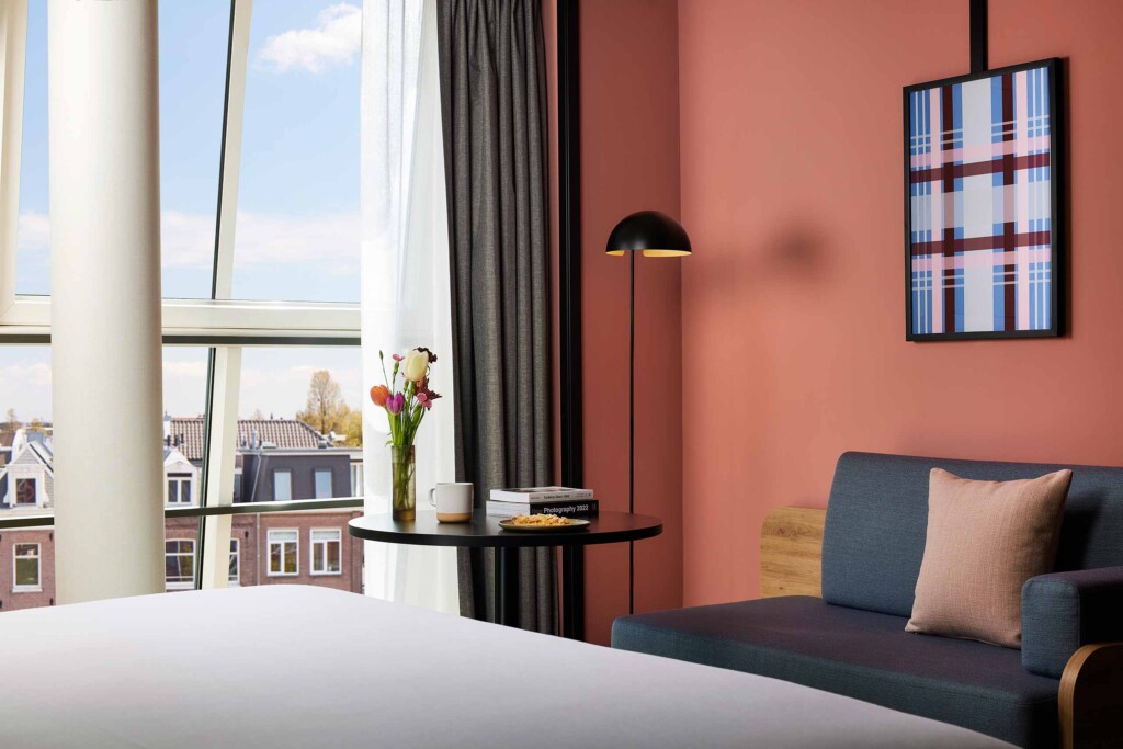View from inside room at Avani Museum Quarter Amsterdam out over the rooftops of the city through chequered glass windows. IN front of the window, an orange armchair with an AvaniFit yoga mat on it, as well as a table with tulips, macaroons and photography artbooks, next to a grey mid-century modern sofa set against a teracotta wall