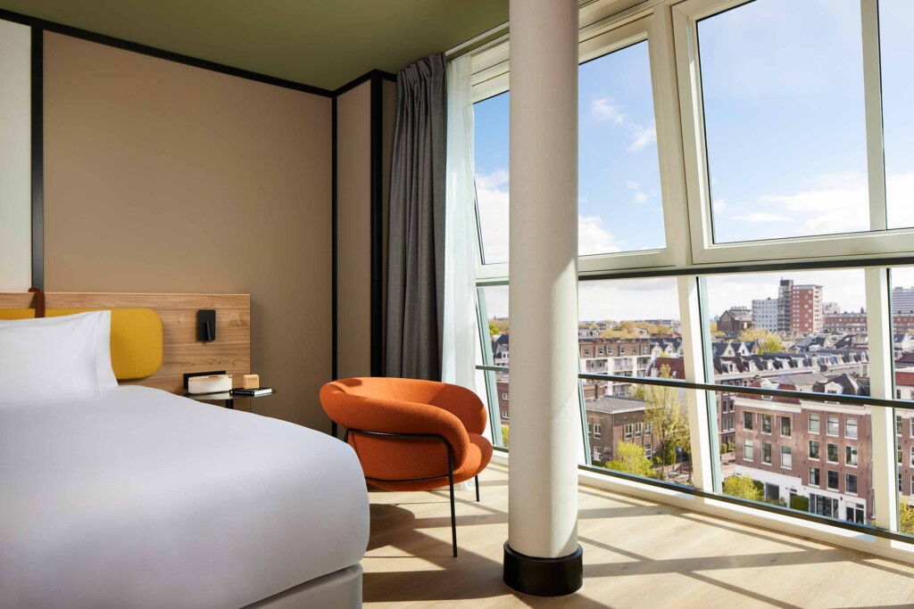 View from inside room at Avani Museum Quarter Amsterdam out over the rooftops of the city through chequered glass windows. In front of the window, an orange armchair and bedside details, with beech wooden floors