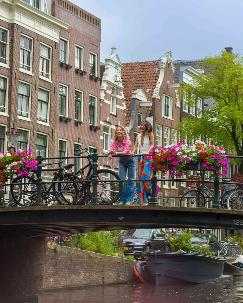 Two women smile and lean over a bicycle laden bridge with flowers, with old Dutch architecture in the background – in Amsterdam. Both are young with long hair, one blonde wearing a pink overshirt and blue jeands, the other wearing a white blouse and pink and blue skirt