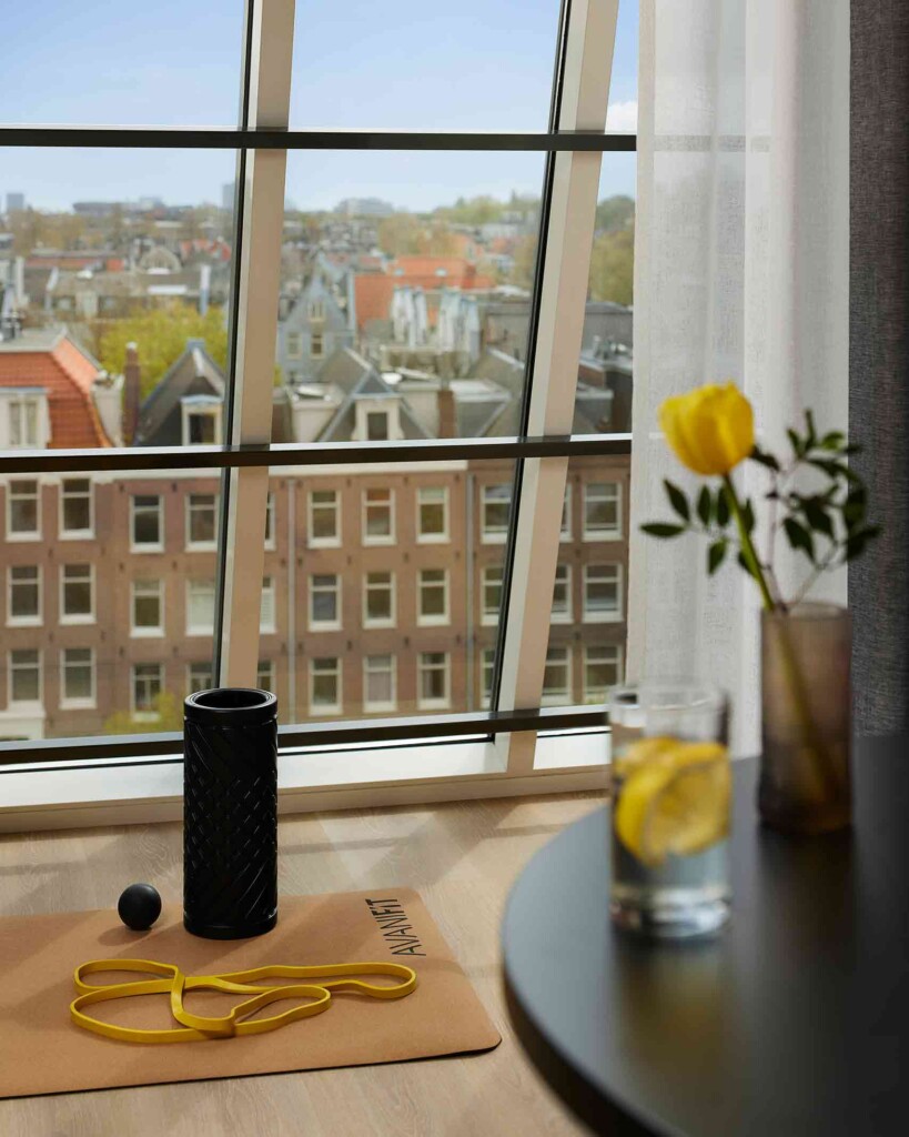 Avani fit yoga mat, with yellow gym band, black roller and massage ball set up looking out the chequered windows of Avani Museum Quarter Amsterdam. In the foreground, out of focus is a glass of water with a lemon in it, as well as a brown glass vase with a single yellow tulip