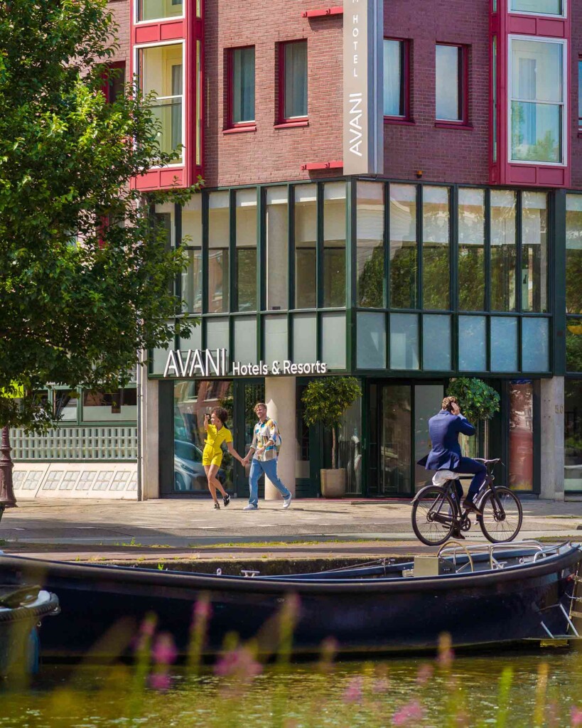 Exterior of the front of Avani Museum Quarter Amsterdam. A businessman in a blue suit rides along the canal where there is an old boat. A young, male-femal couple skip hand in hand – she is black with curly hair and wears a yellow romper. He is white, with blonde curly hair and wears a colourful shirt and blue jeans