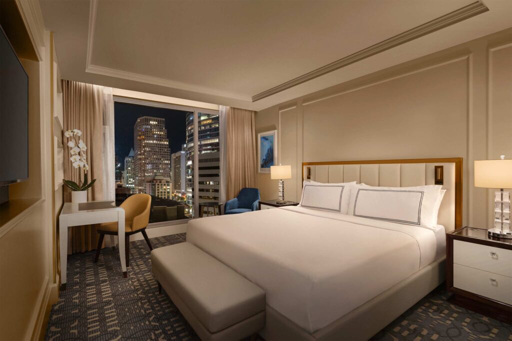 A king-size bed dressed in white bedlinen, with a bedside table lamp on each side and a small white desk in front of it, inside a warmly lit suite at AZUR Legacy Collection Hotel, Vancouver, Canada