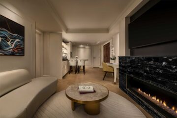 A curved couch across from a gas fireplace with a large flatscreen TV above it inside a beige, moodily lit suite at AZUR Legacy Collection Hotel, Vancouver, Canada