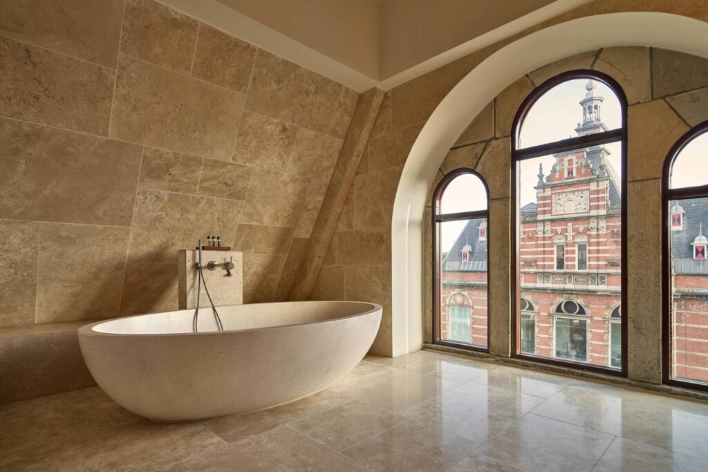 Bathroom at Mandarin Oriental Conservatorium Amsterdam in the rooftop, windows looking out to another, red-brick heritage building. This bathroom is very minimalist with gold marble tiling and nothing but a stone bath sat in the space