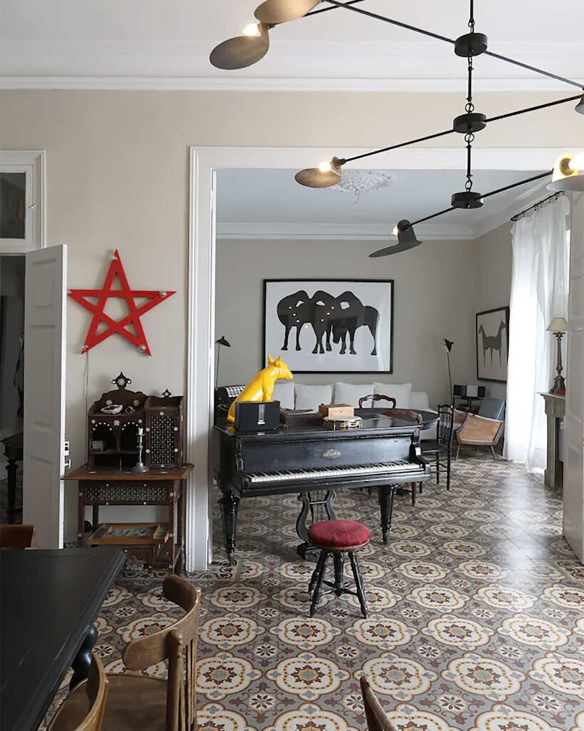 An airy living room at Mimi Calpe with patterned tiled floors features a black grand piano, eclectic furniture, and contemporary artwork, with soft daylight filtering through tall windows.