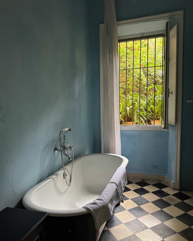 A simple bathroom at Mimi Calpe with blue-painted walls and a freestanding bathtub sits beside an open window, looking out onto leafy garden foliage.