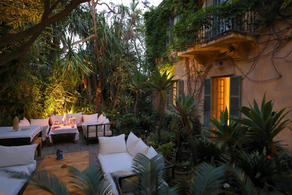 A garden at dusk is softly lit with warm lights, showing outdoor dining tables and cushioned seating surrounded by dense greenery and climbing plants.