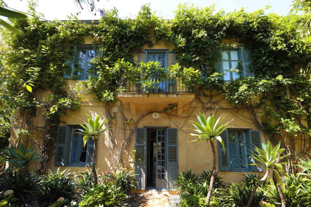 A pale yellow villa façade is partially covered in climbing vines, with green shutters, small balconies, and dense planting framing the entrance.