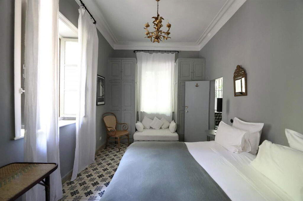 A calm, light-filled bedroom at Mimi Calpe with grey walls, high ceilings, and patterned tiles features a neatly made bed, soft curtains, and a small seating area.