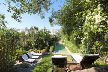 A lush, terraced garden with dense greenery surrounds a long, narrow swimming pool, with sun loungers set on a stone patio overlooking the city beyond.